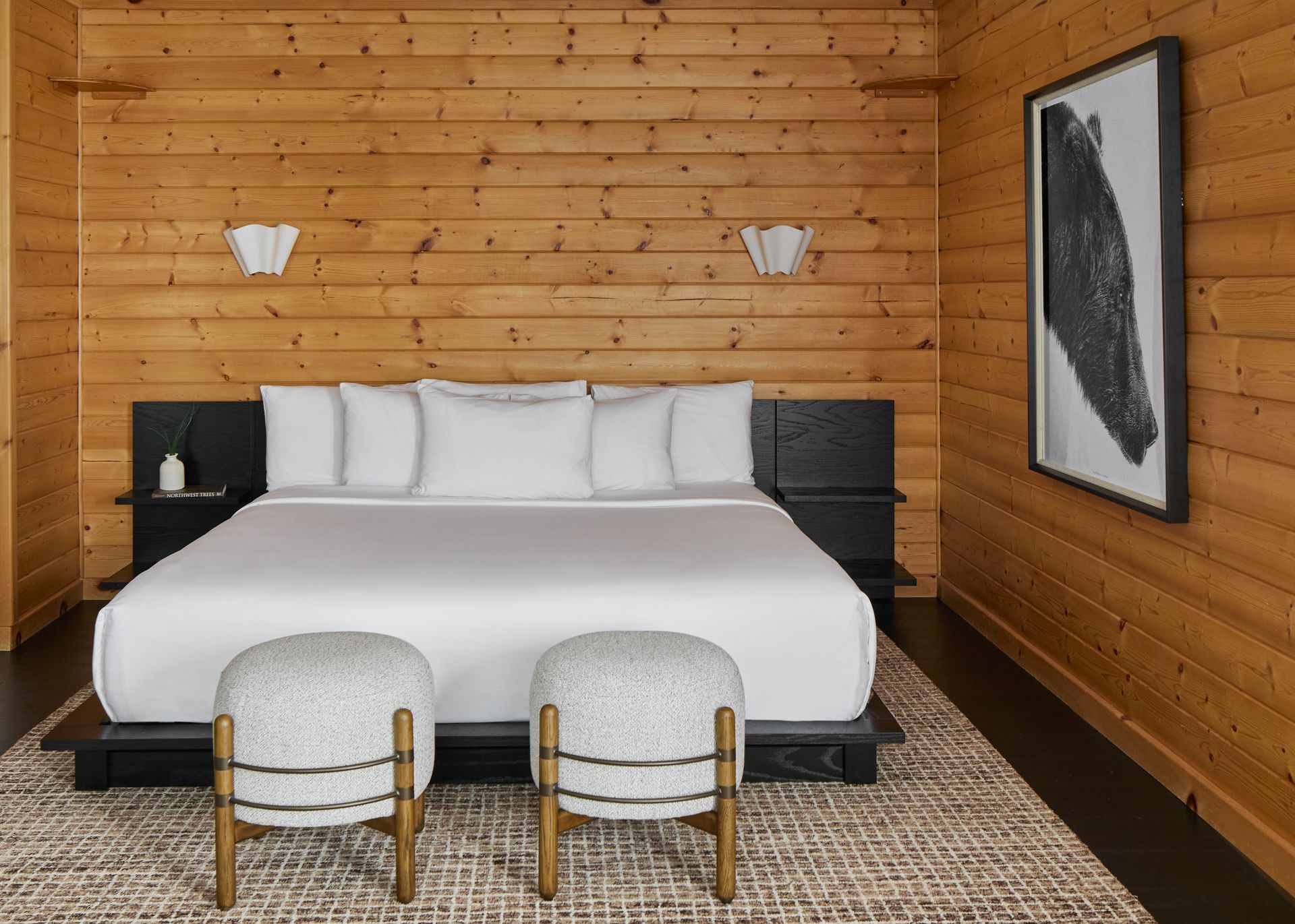 Cozy wooden bedroom with a white bed, two stools, wall sconces, and framed art.