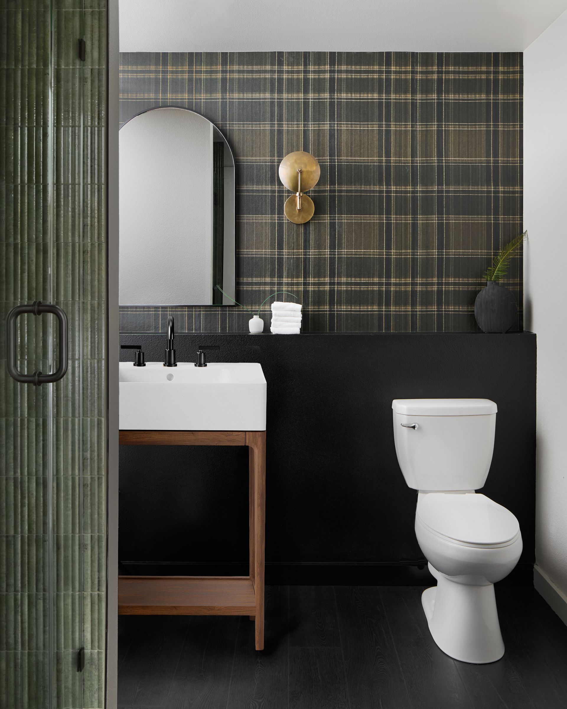 Modern bathroom with black tiled wall, white toilet, sink, mirror, and gold wall sconce