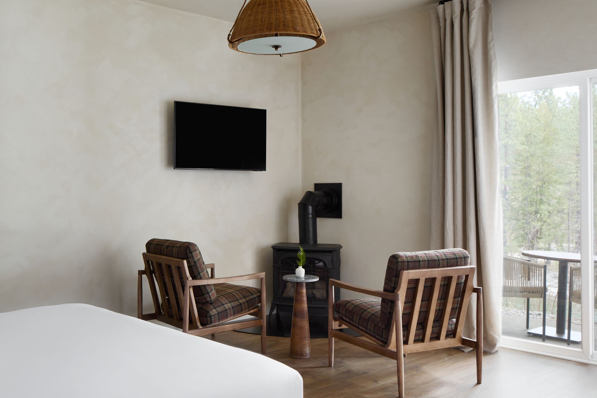 Modern hotel room with two wooden chairs, small table, wall-mounted TV, and balcony door.