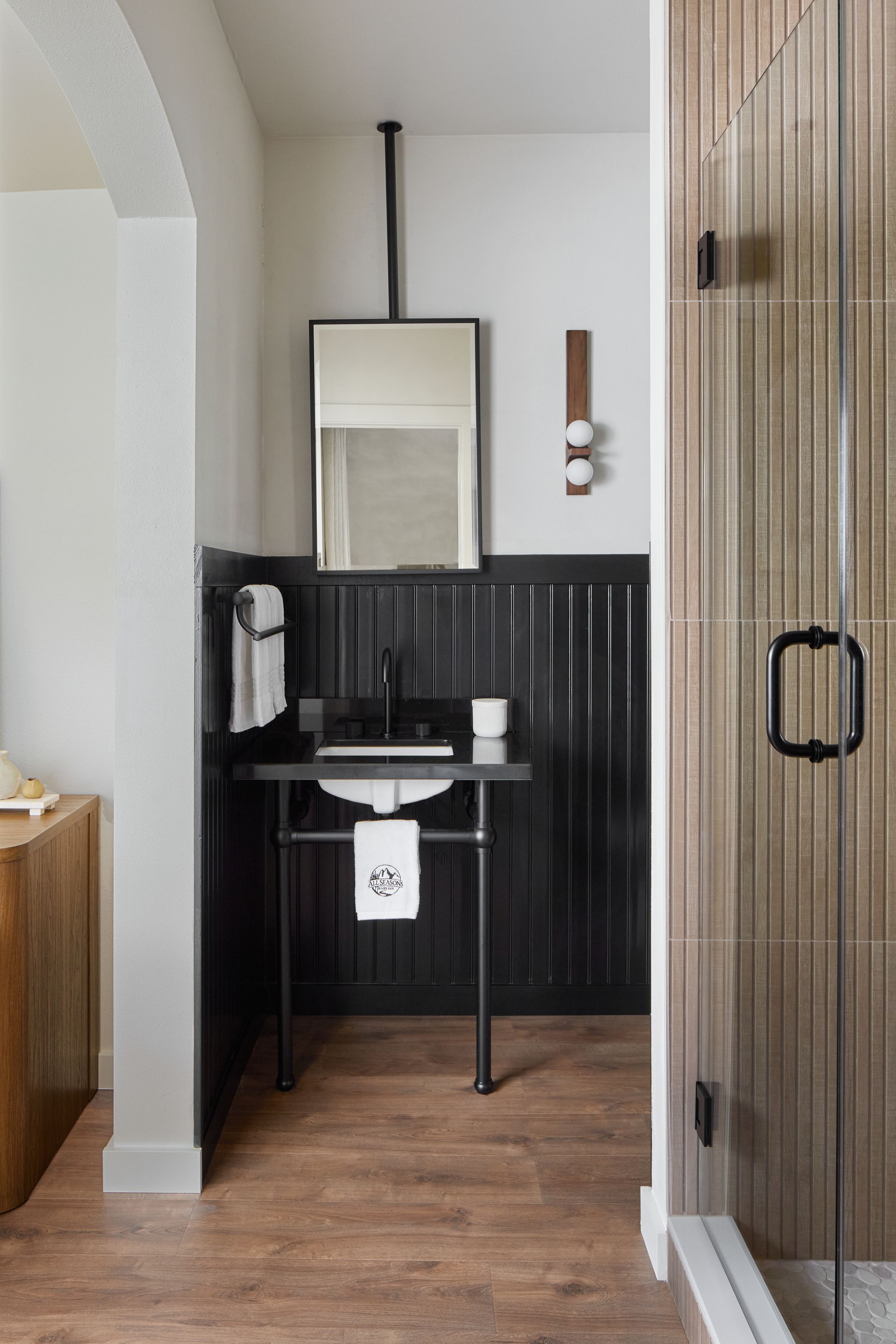 Black pedestal sink and mirror in a narrow bathroom with wood floor and dark wainscoting.