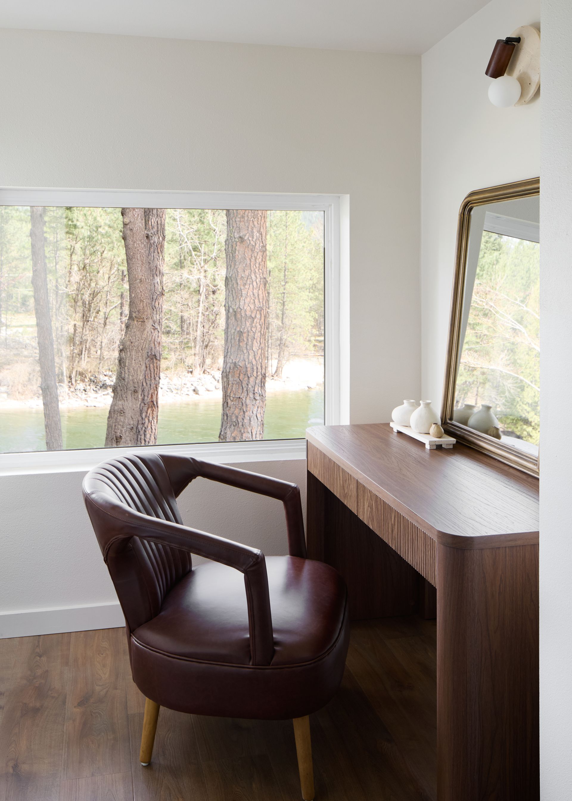 Brown leather chair beside a wooden desk and ornate mirror in a bright room with a large window.