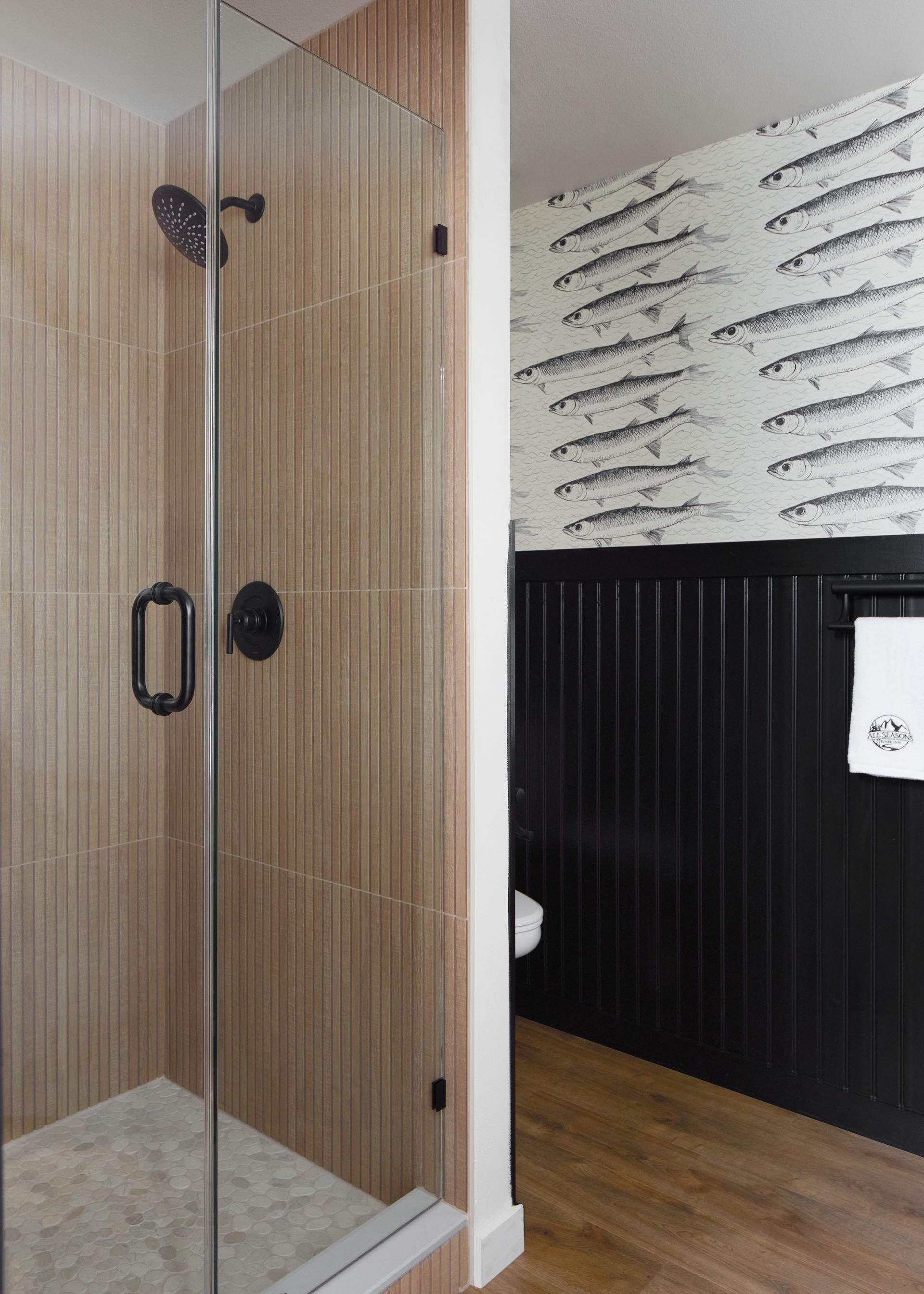 Glass shower enclosure beside black wainscoting and patterned wallpaper on wood flooring.