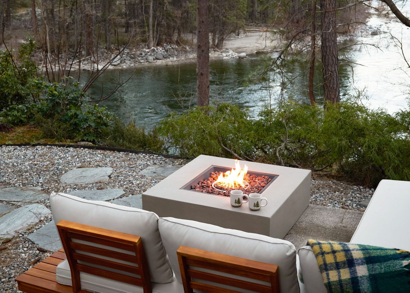 Outdoor patio with a stone fire pit beside a river, surrounded by chairs and greenery