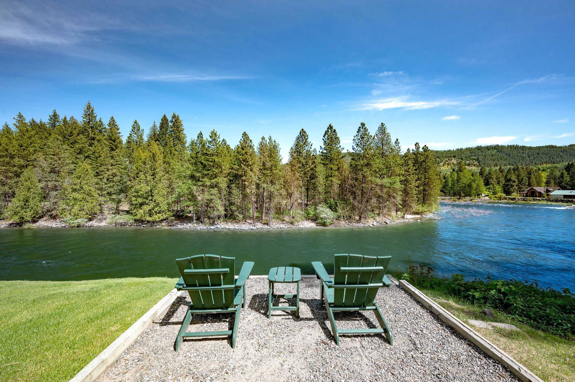 Two green chairs on the Wenatchee river facing calm water and pine trees under a blue sky