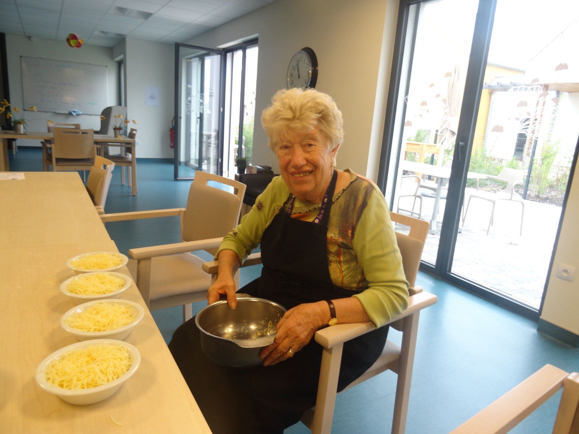 Une femme âgée est assise à une table tenant un bol de nourriture.