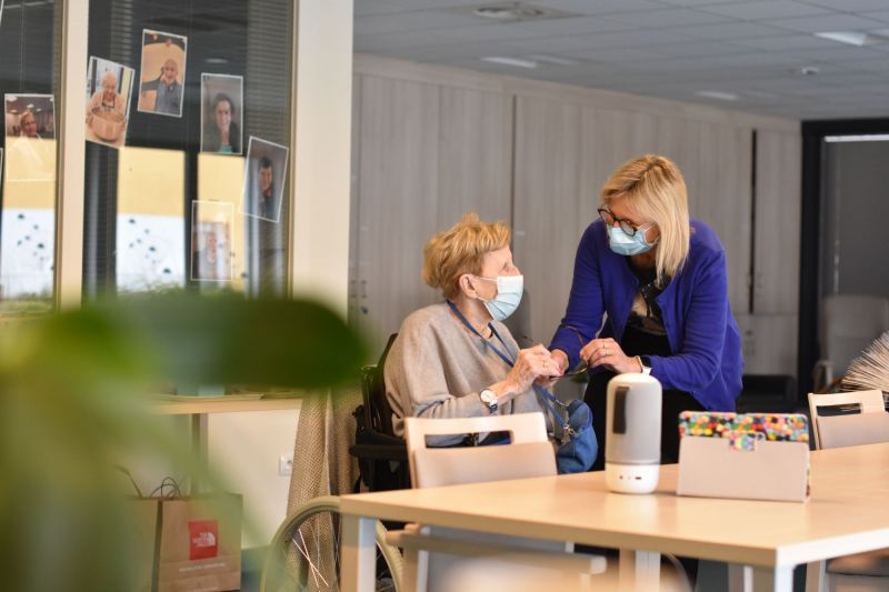 Une femme portant un masque parle à une femme âgée assise à une table.