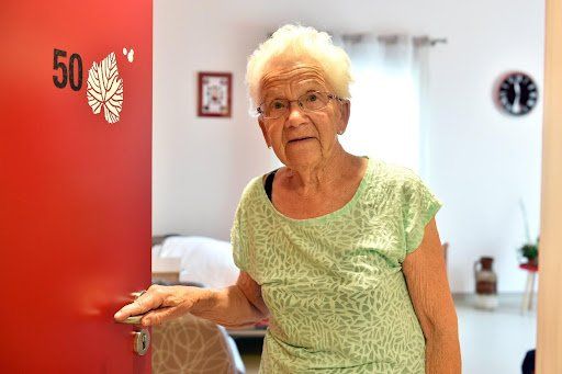 Une femme âgée se tient devant une porte rouge.