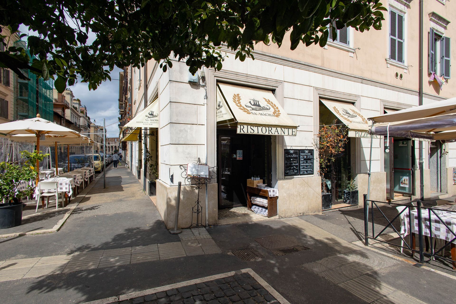 Ingresso del caffè che si affaccia sulla strada, con tende da sole color crema, tavolini all'aperto e un albero che proietta ombra sul marciapiede.