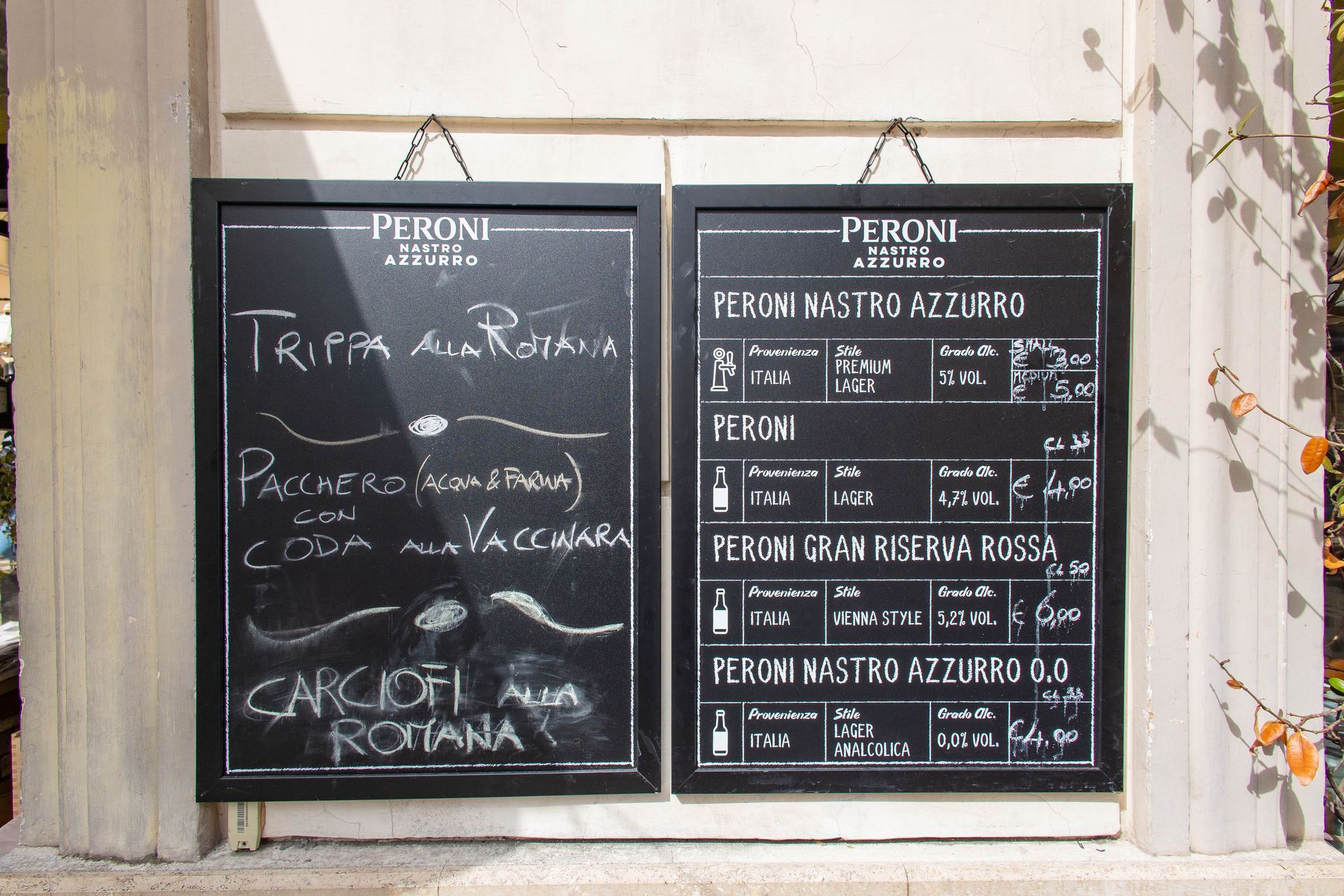 Due menù di ristorante su lavagna nera in una cornice bianca, con testo italiano scritto a mano su un muro di pietra.