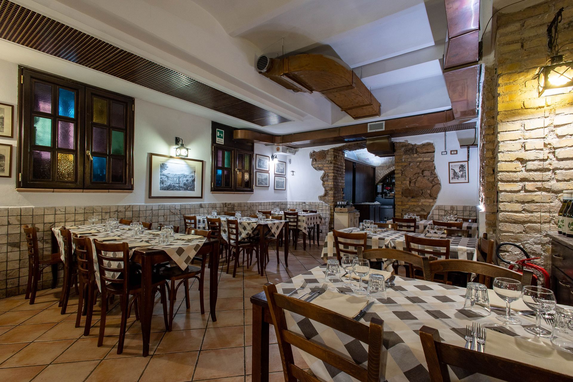 Accogliente interno rustico di un ristorante con tovaglie a quadri, sedie in legno e pareti in pietra illuminate da una luce calda.