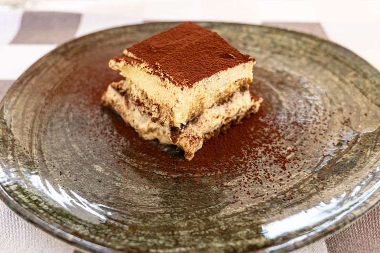 Un piccolo tiramisù quadrato spolverato di cacao su un piatto di ceramica marrone.