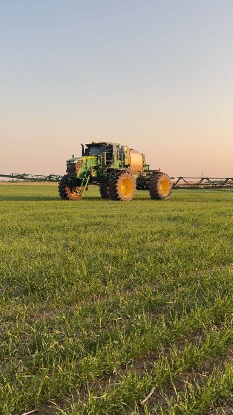 A green and yellow tractor sprays a field of green crops under a soft, golden sunset.