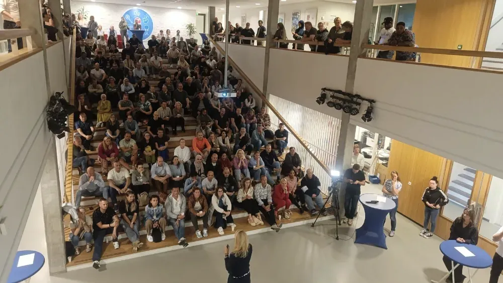 Een vrouw spreekt in een microfoon voor een zittend publiek in een lichte, moderne zaal met oplopende zitplaatsen en planten.