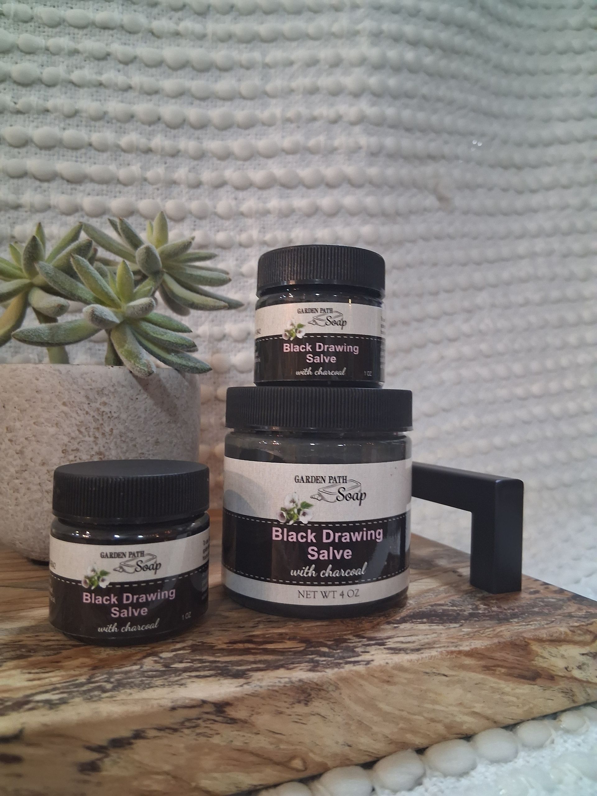 Four jars of Black Drawing Salve stacked on a wooden board next to a small succulent plant.