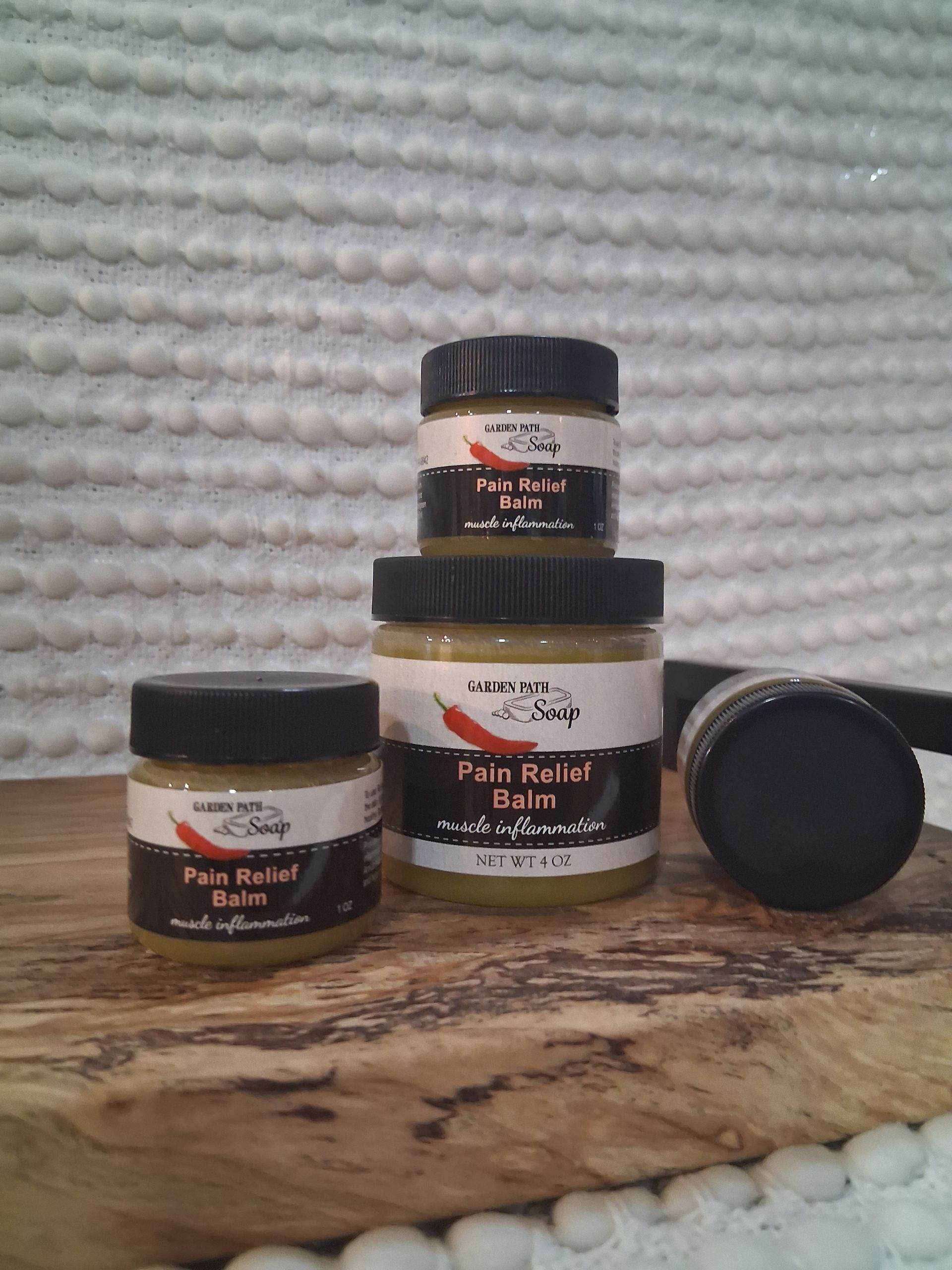 Four jars of pain relief balm, stacked on wooden surface against white textured background.