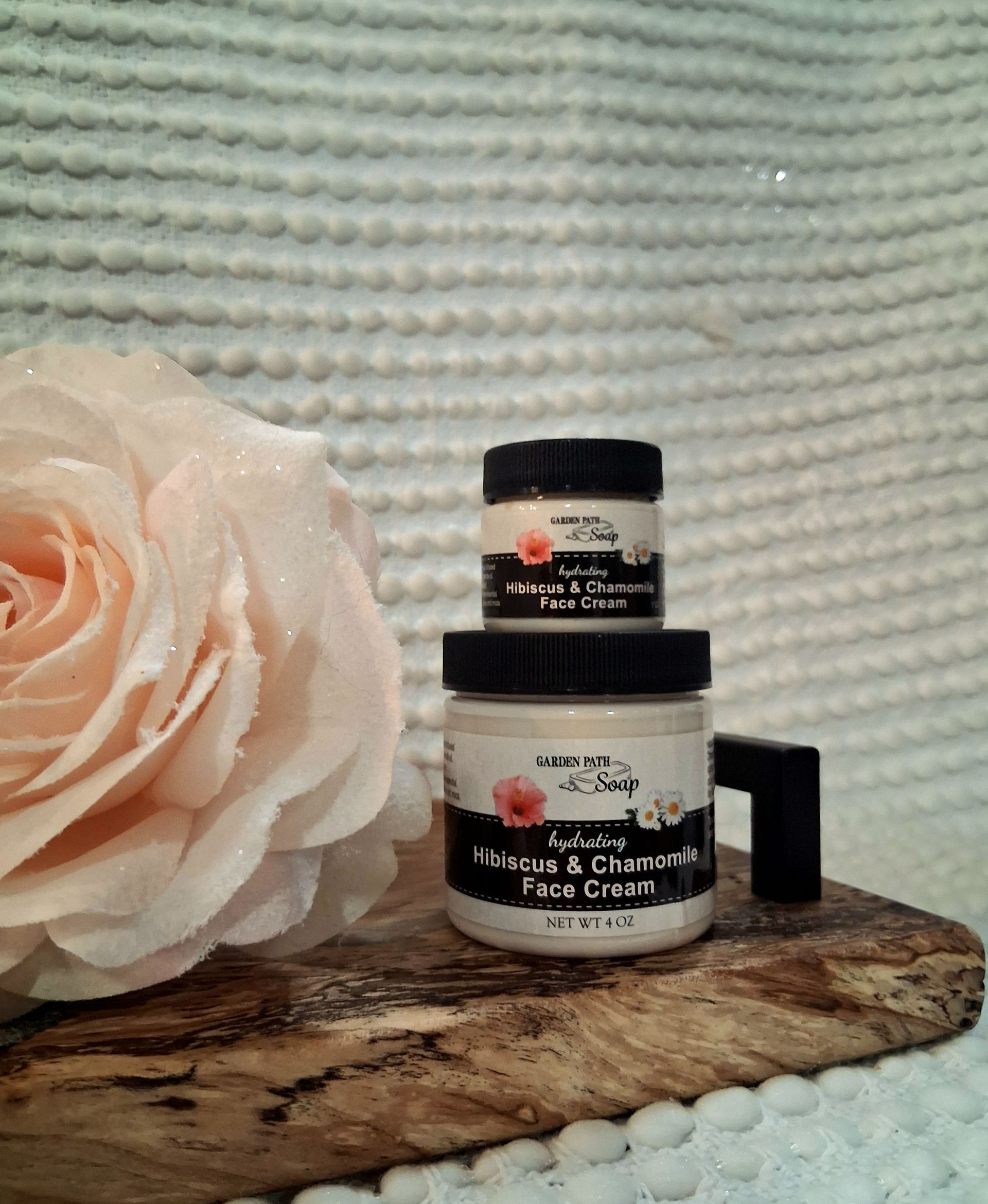 Two cream jars stacked, with a rose, on a wooden surface, against a textured white background.