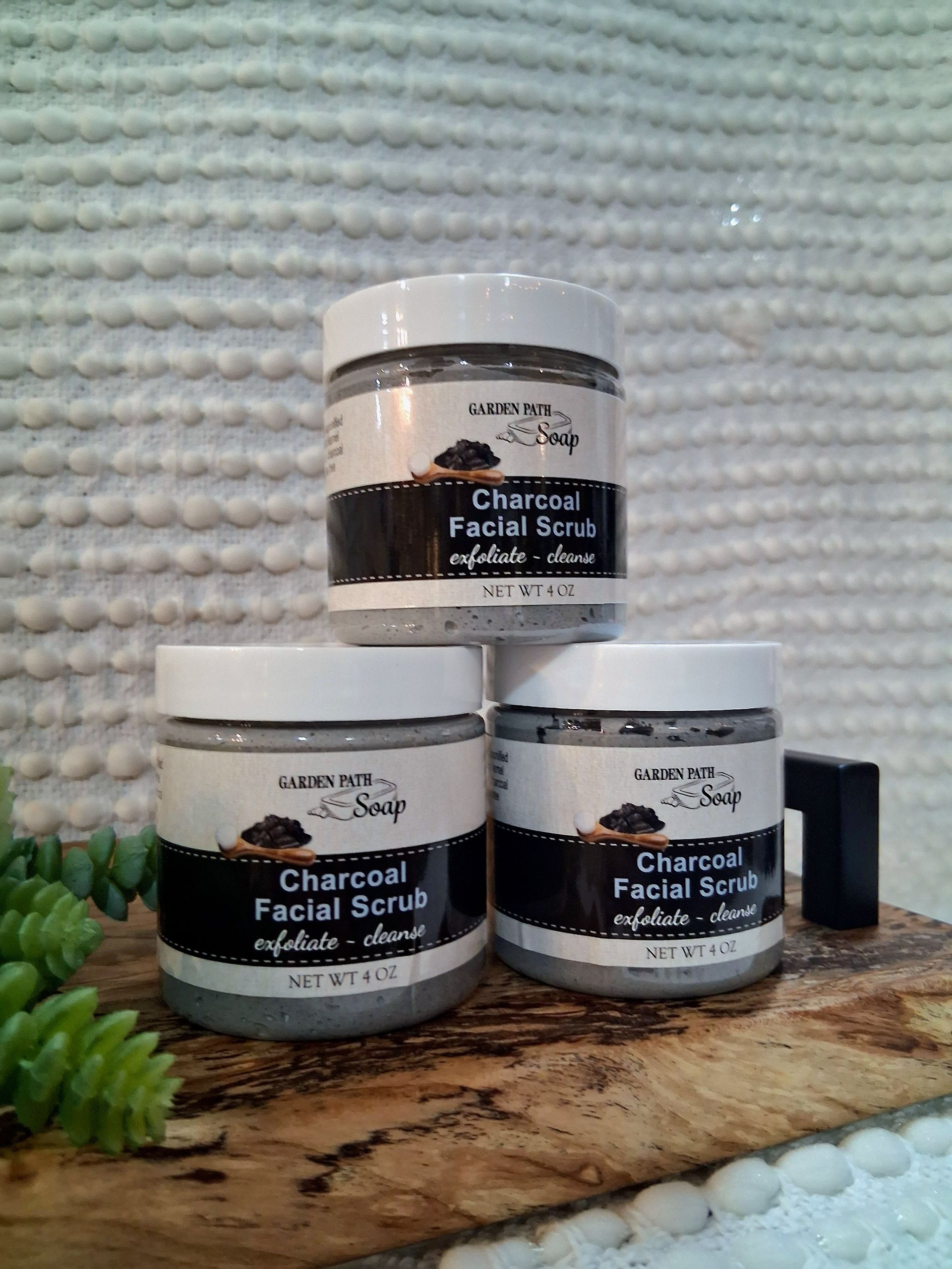 Three jars of charcoal facial scrub stacked on a wooden surface, with greenery in the background.