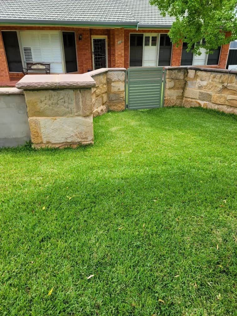 A Lush Green Lawn In Front Of A Brick House With A Stone Wall — Anne & Ron Johnson Fencing in Werris Creek, NSW