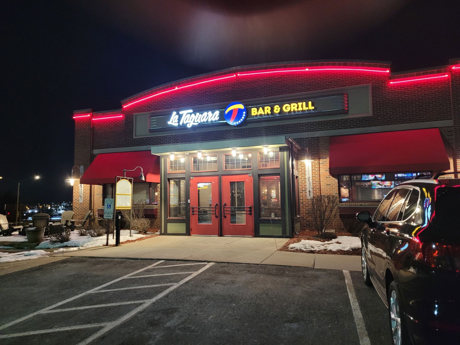 A lit bar and grill storefront at night with red trim and parked cars in front.