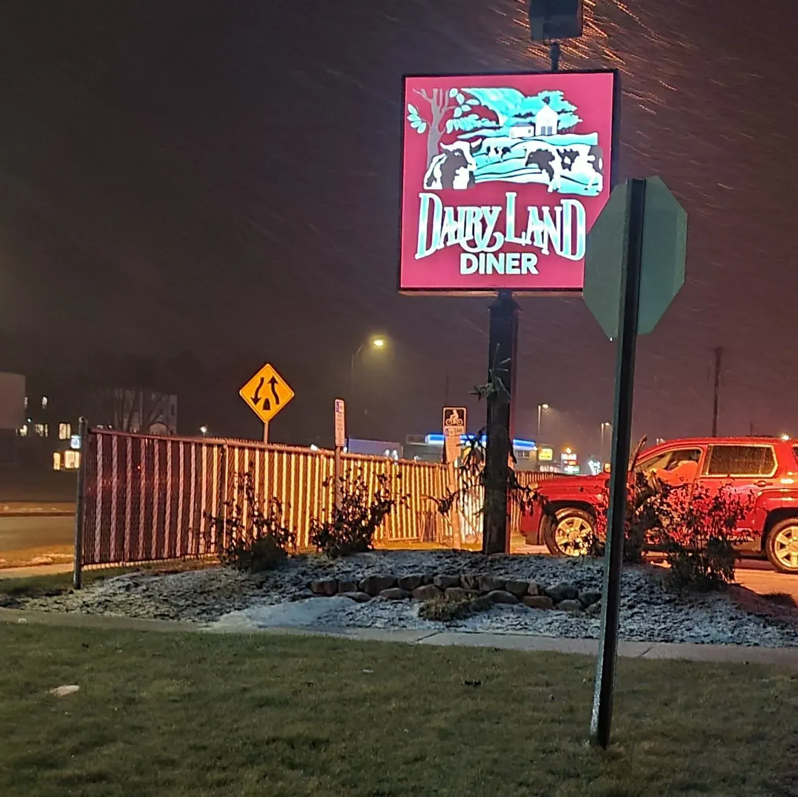 Night view of a lit “Dairyland Diner” sign above a roadside parking lot with cars and a fence.