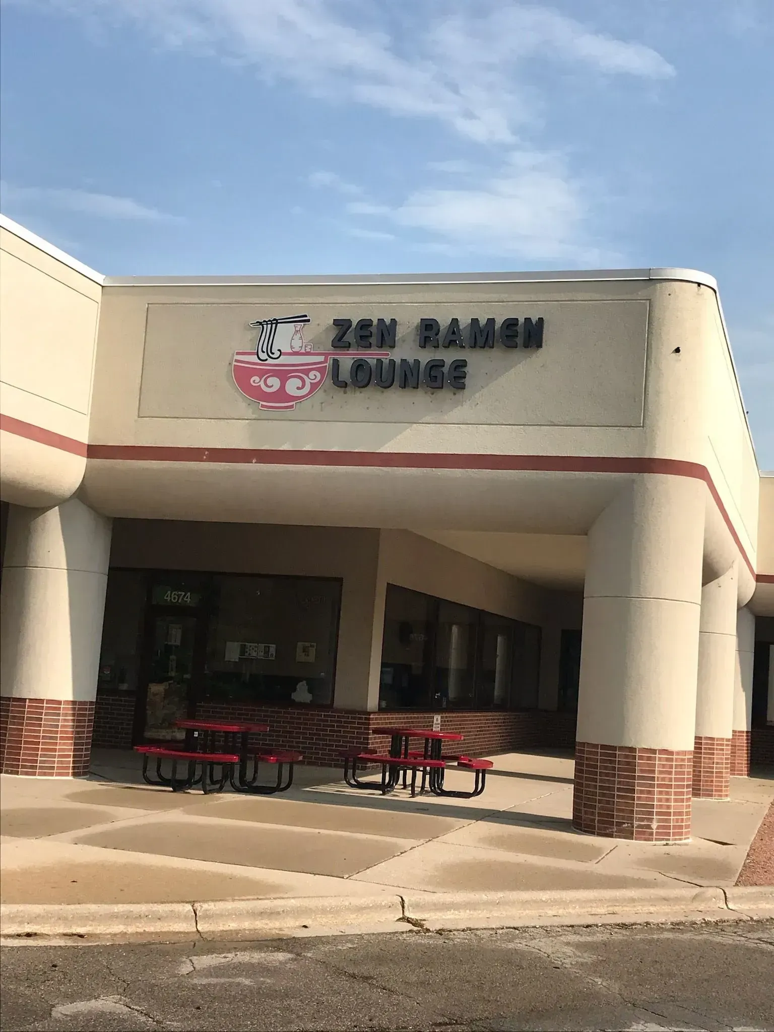 Storefront entrance with Korean sign, tan facade, and outdoor picnic tables under a covered walkway.