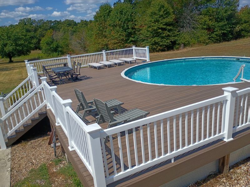 Wooden pool deck with white railings, lounge chairs, and a blue swimming pool in a sunny yard.