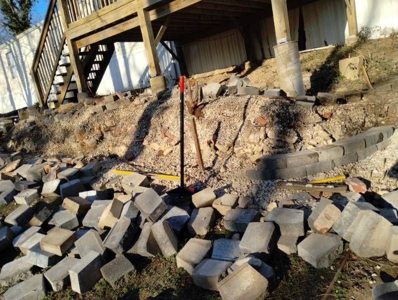 Damaged hillside beneath a building, with collapsed retaining wall blocks and exposed soil.