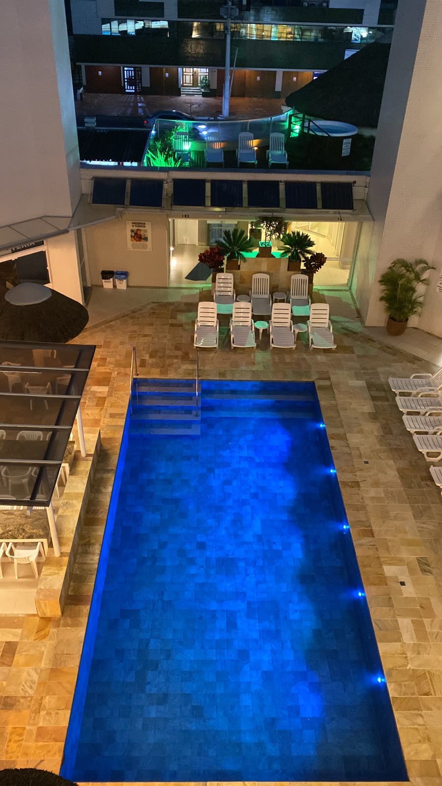 Pátio de hotel à noite, com uma piscina azul iluminada, espreguiçadeiras e varandas nos edifícios ao redor.