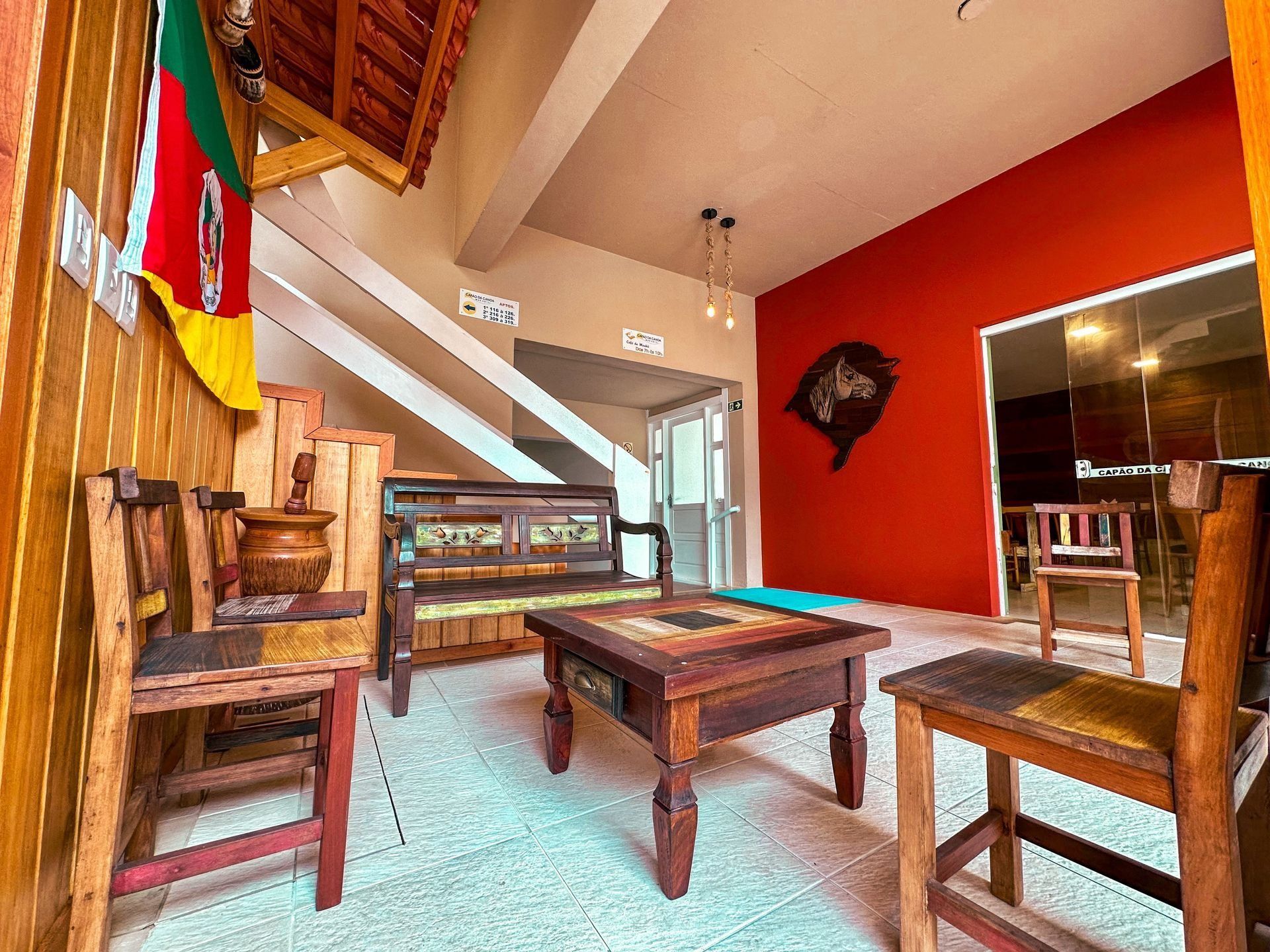 Sala de estar rústica com móveis de madeira, parede de destaque vermelha, escada e janelas amplas