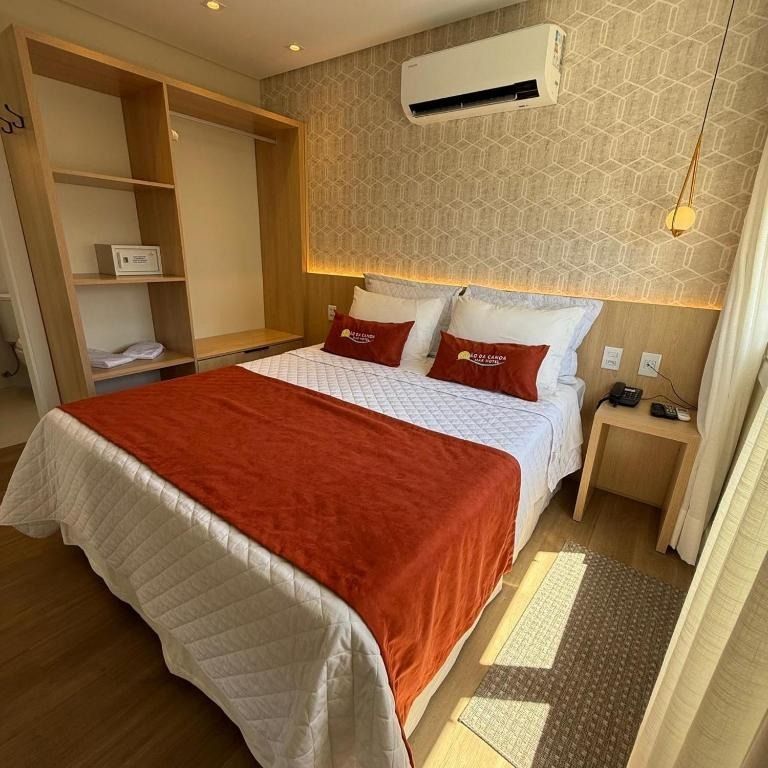 Quarto de hotel arrumado, com roupa de cama vermelha, prateleiras de madeira, mesa de cabeceira e ar-condicionado de parede