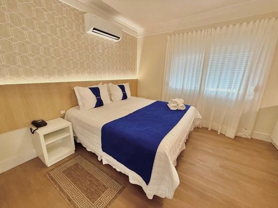 Quarto iluminado, com a cama bem-arrumada, detalhes em azul, cortinas brancas e piso de madeira clara.