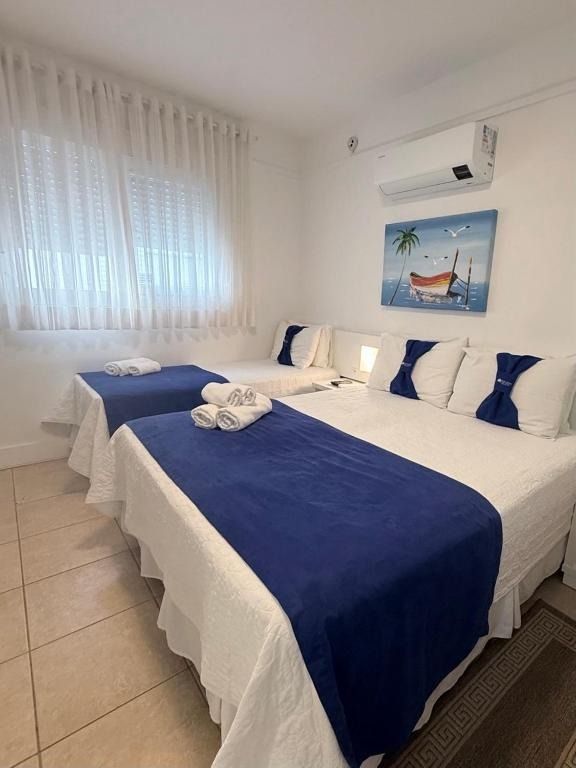 Quarto luminoso com duas camas de solteiro vestidas com roupa de cama azul e branca, piso de azulejos, janela e quadros decorativos.