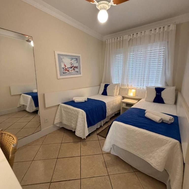 Quarto luminoso com duas camas de solteiro, roupa de cama azul, um abajur, espelho e piso de ladrilhos