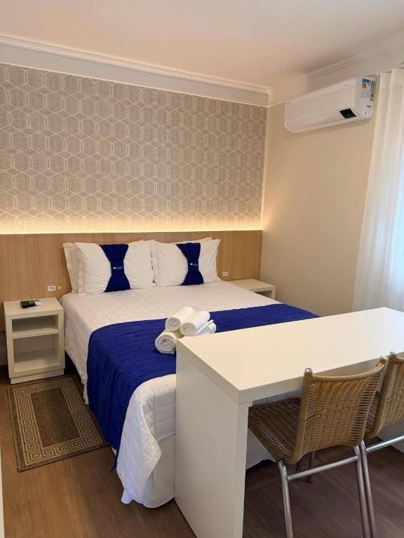 Quarto compacto com cama azul e branca, secretária e cadeiras, ar condicionado de parede e iluminação suave