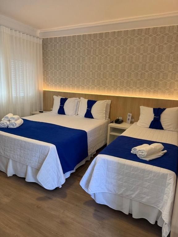 Quarto de hotel com duas camas de solteiro, roupa de cama branca, peseiras azuis e toalhas dobradas aos pés de cada cama