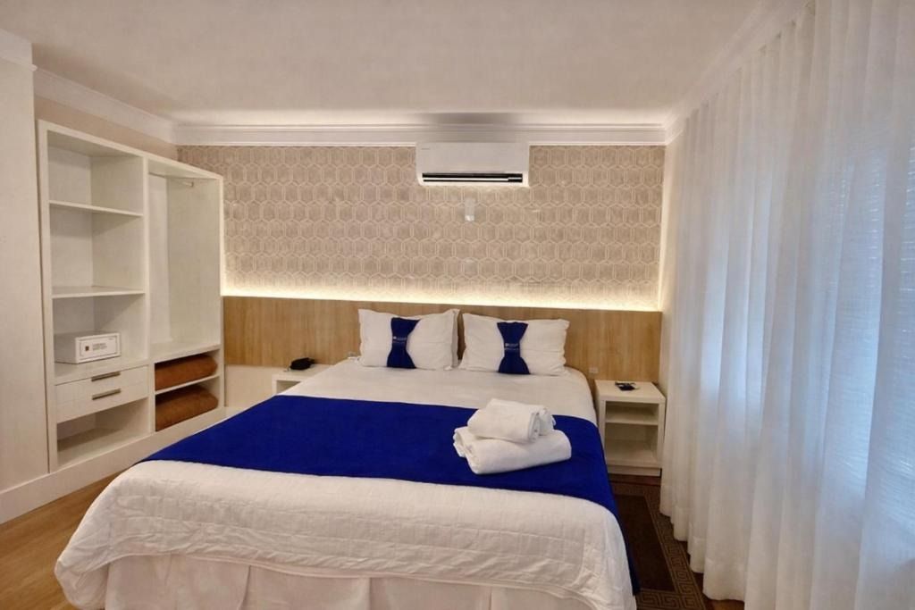 Quarto bem arrumado, com roupa de cama azul, prateleiras brancas e cortinas transparentes junto à janela