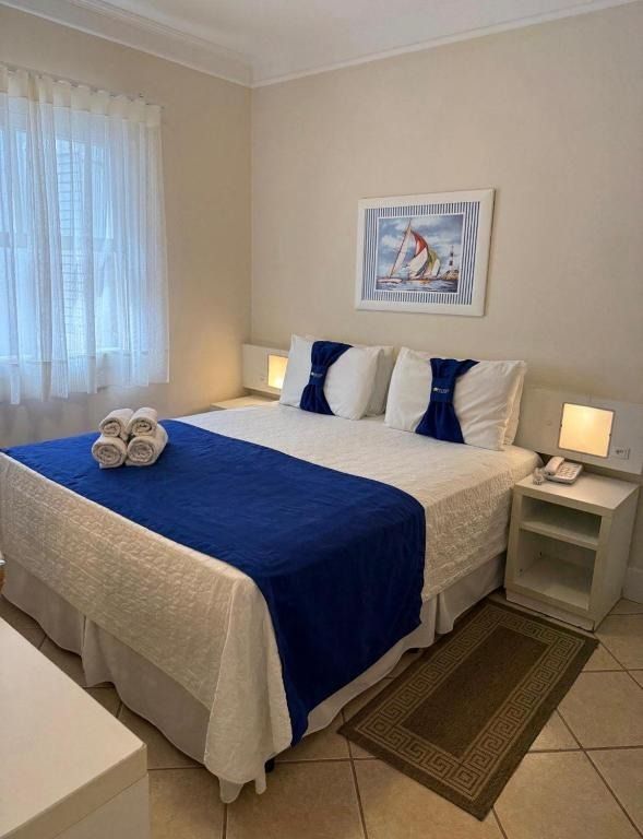 Quarto aconchegante com cama azul e branca, candeeiros de cabeceira, janela e quadros emoldurados na parede