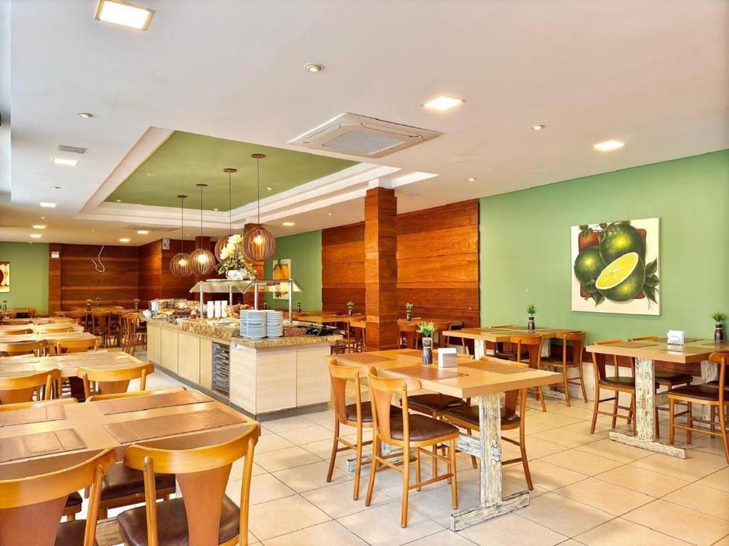 Área de refeições iluminada de um café, com mesas de madeira, bancos, piso de ladrilhos e detalhes verdes nas paredes