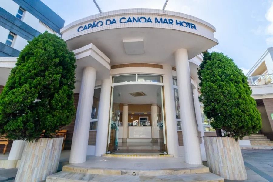 Entrada de um edifício moderno com colunas brancas, portas de vidro e arbustos verdes aparados sob uma placa curva.