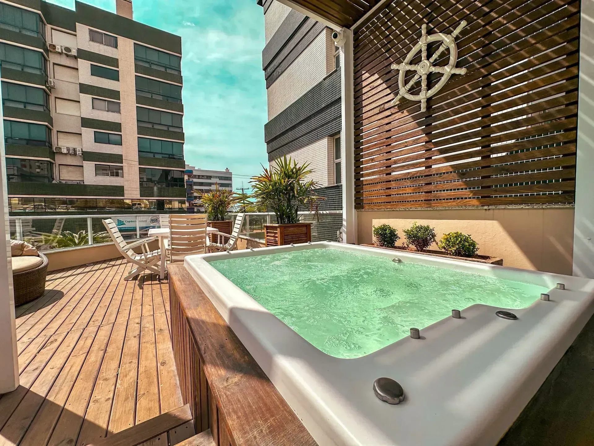 Terraço na cobertura com uma banheira de hidromassagem branca, piso de madeira, espreguiçadeiras e edifícios da cidade ao fundo