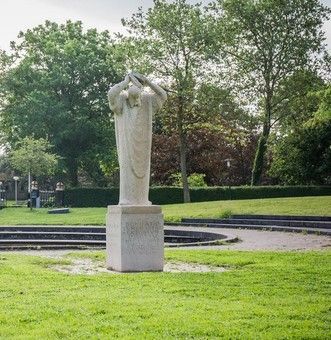 Standbeeld in een park. Een lange, gedrapeerde figuur op een sokkel. Groen gras, bomen en cirkelvormige, trapsgewijs opgestelde structuren op de achtergrond.