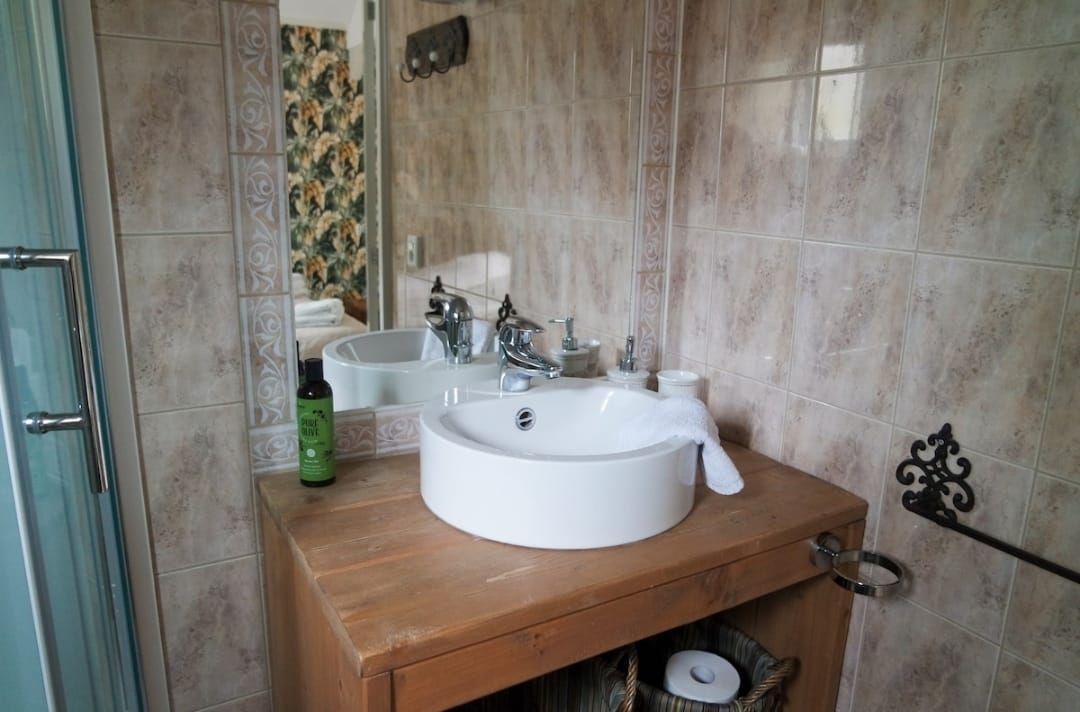 Badkamer met een witte wastafel op een houten meubel, een grote spiegel, beige betegelde muren en een glazen douchedeur.