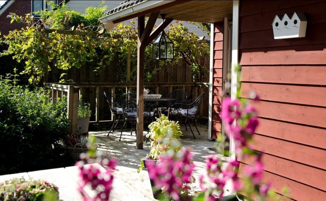 Een rood gebouw met een kleine houten veranda, terrasmeubilair en roze bloemen op de voorgrond.