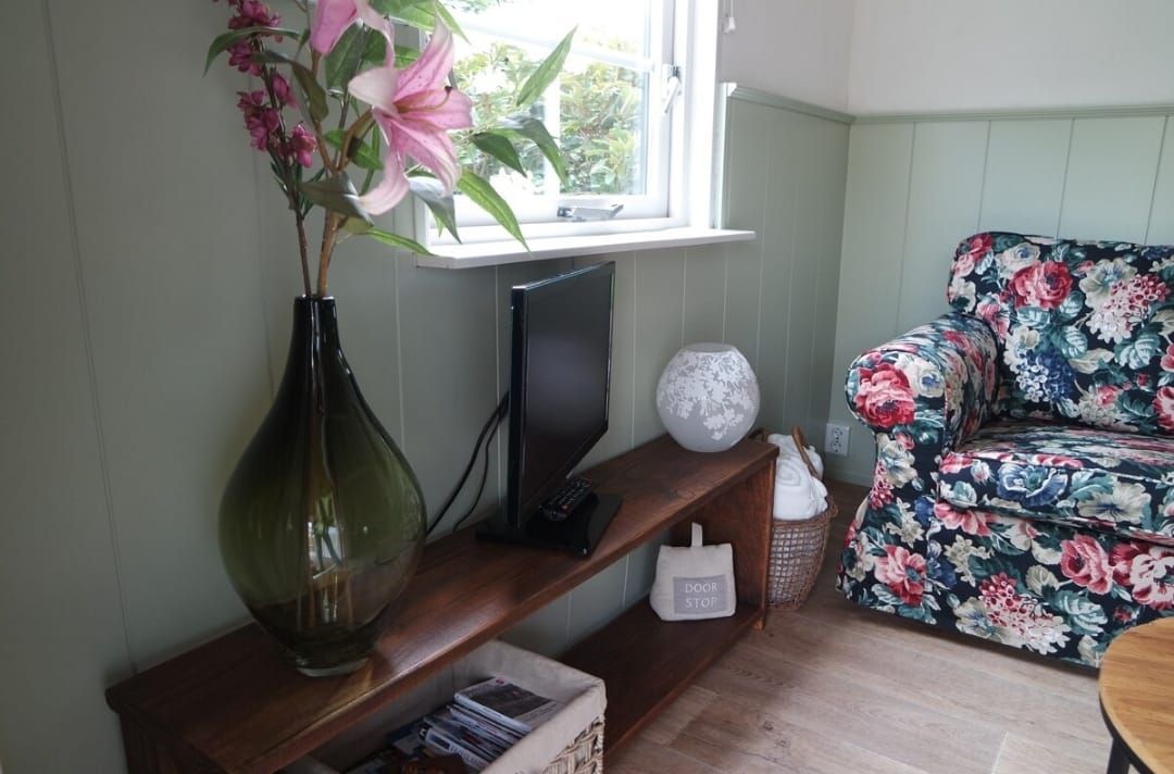 Een knusse kamer met een fauteuil met bloemenprint, een tv en bloemen in een grote groene vaas.