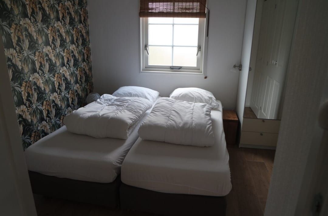 Twee eenpersoonsbedden met wit beddengoed in een kamer met behang met een patroon, een raam en een klein nachtkastje.