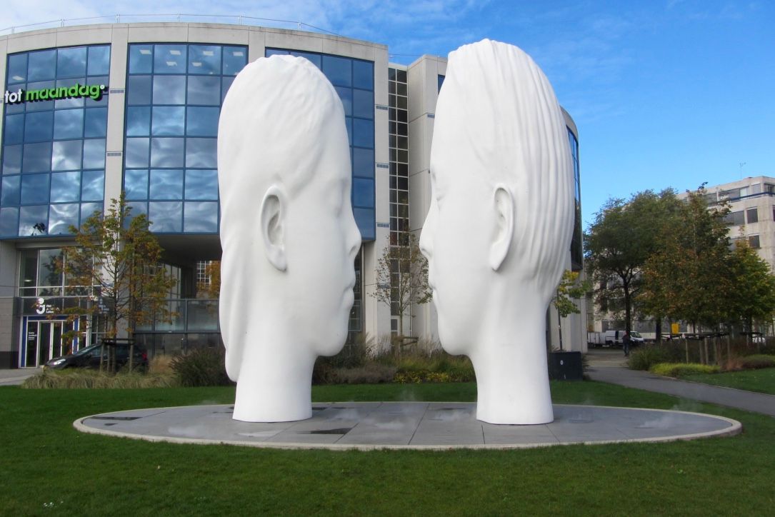 Twee grote, witte sculptuurhoofden staan ​​tegenover elkaar, voor een modern gebouw met groene bewegwijzering en een grasveld.