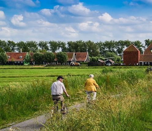 Een stel fietst over een pad door een veld, met traditionele Nederlandse huizen op de achtergrond.