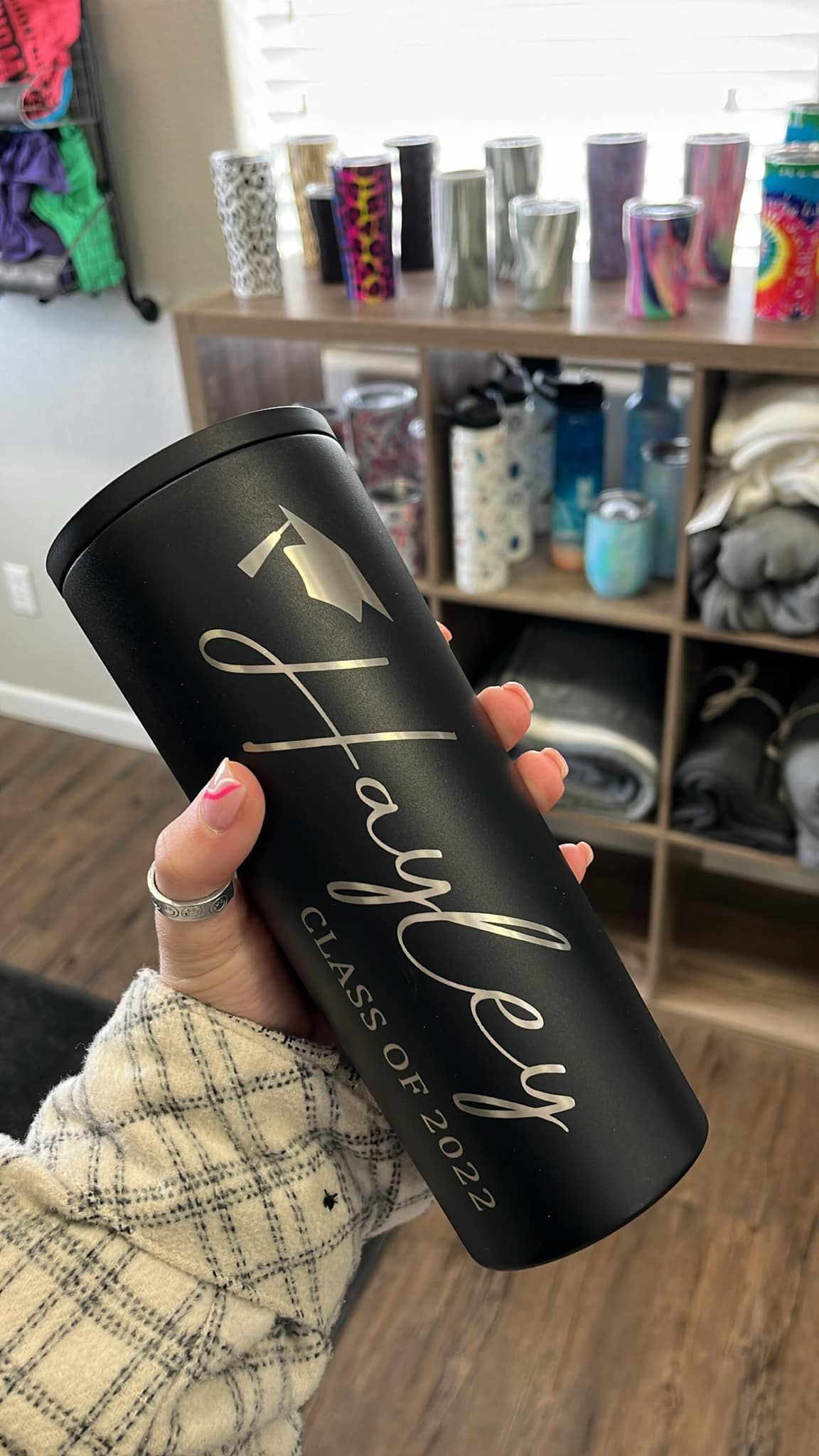 Hand holding a black tumbler with gold script in front of a shelf of drink cups and bottles