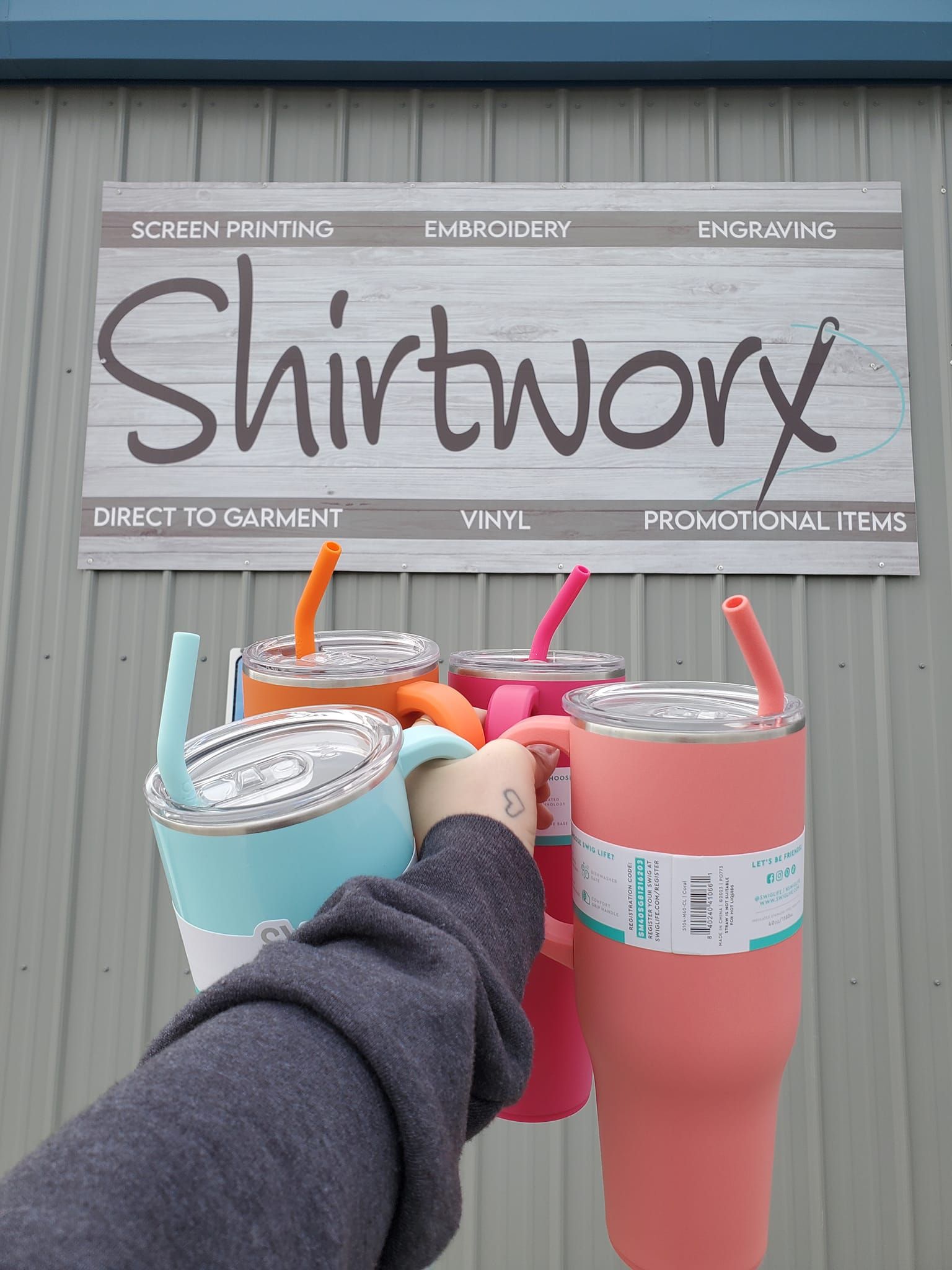 Three colorful drinks held up in front of a Shirttory sign outdoors