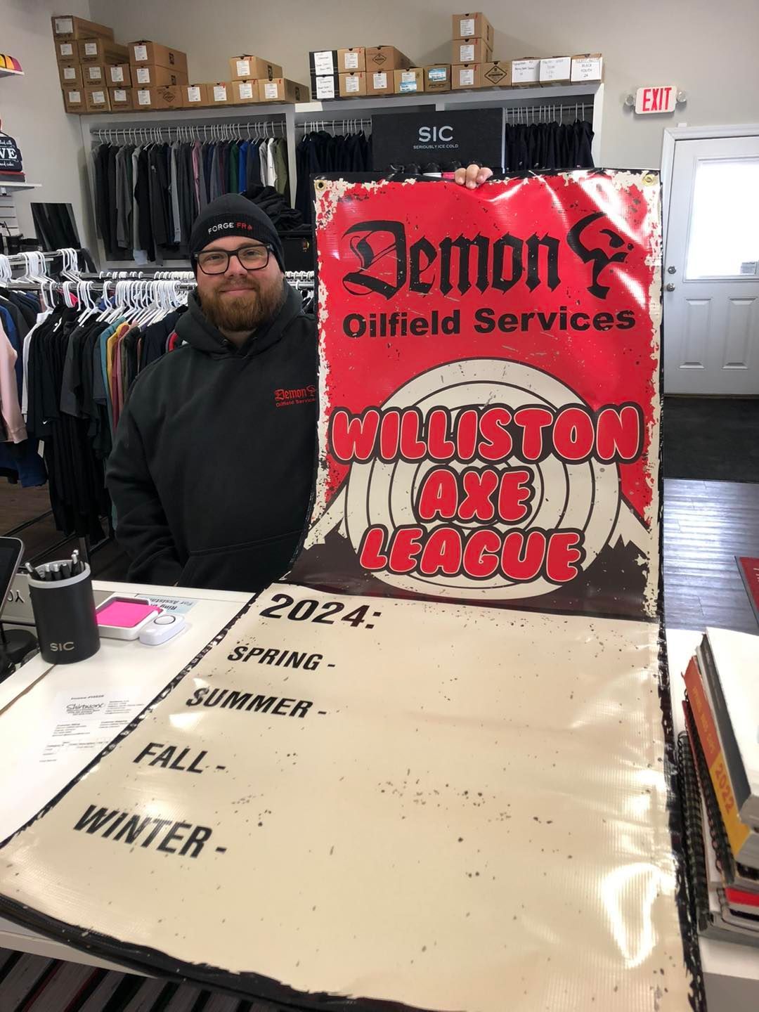 Demon Oilfield Services employee beside a “Williston Axe League” sign in a clothing store, with fall and winter listings.